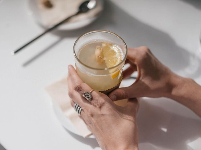 Die Hände einer Frau halten auf einem Tisch ein Glas mit frischem Ingwertee fest. | © gettyimages.de / Westend61