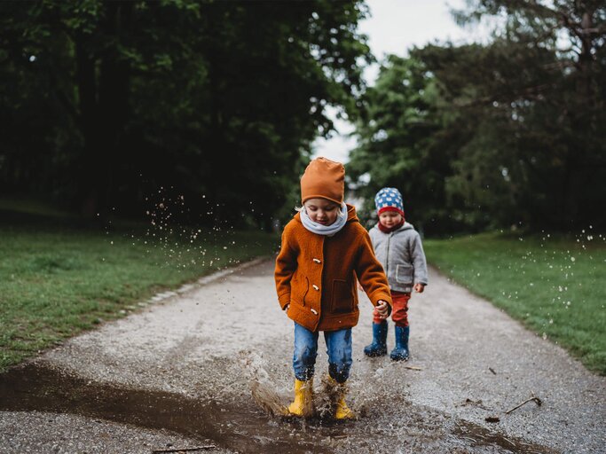 Zwei Kinder spielen an der frischen Luft, ein Kind springt mit Gummistiefeln in eine Pfütze. | © gettyimages.de / Cavan Images