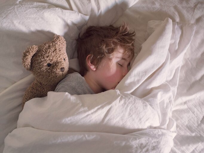 Kind liegt mit einem braunen Teddybär im Bett und schläft. | © gettyimages.de / Christopher Hopefitch