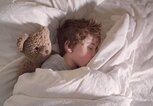 Kind liegt mit einem braunen Teddybär im Bett und schläft. | © gettyimages.de / Christopher Hopefitch