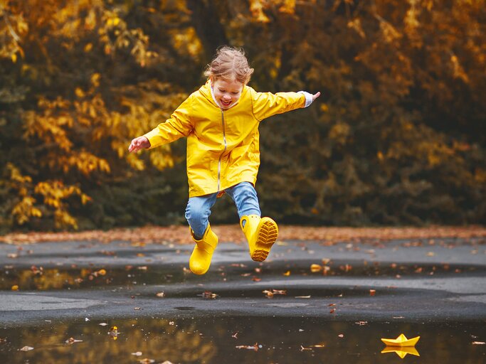 Junges Mädchen mit einer gelben Regenjacke und gelben Gummistiefeln springt im Herbst in eine große Pfütze. | © gettyimages.de / evgenyatamanenko