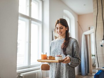 Junge Frau läuft mit einem Frühstückstablett durch ihr Wohnzimmer.  | © Getty Images / Luis Alvarez