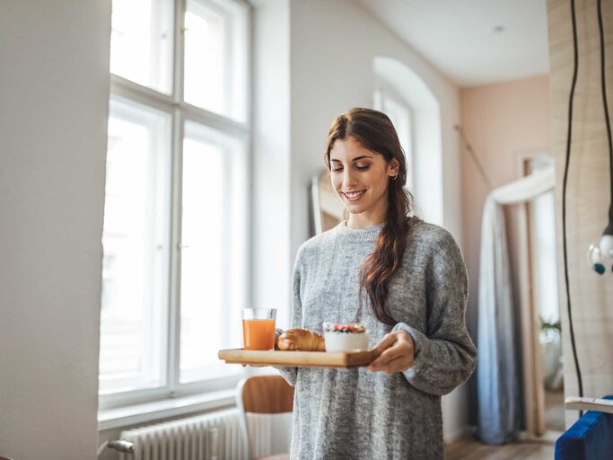 Junge Frau läuft mit einem Frühstückstablett durch ihr Wohnzimmer.  | © Getty Images / Luis Alvarez