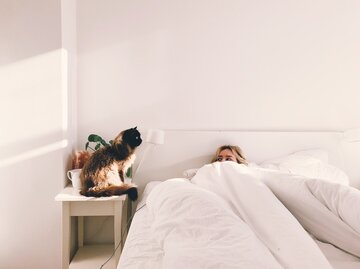 Frau liegt im Schlafzimmer im Bett und neben ihr sitzt eine Katze | © Getty Images/	Adam Kuylenstierna / EyeEm
