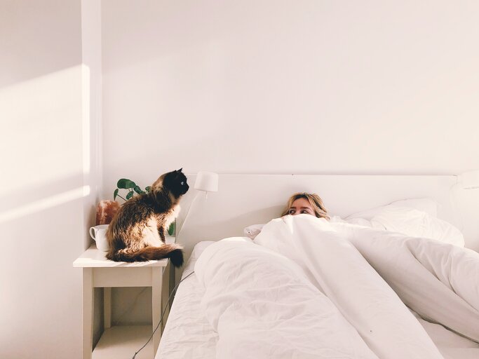 Frau liegt im Schlafzimmer im Bett und neben ihr sitzt eine Katze | © Getty Images/	Adam Kuylenstierna / EyeEm