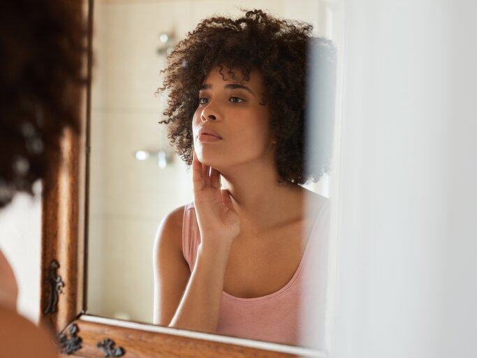 Junge Frau blickt prüfend in den Spiegel | © GettyImages/Mavocado
