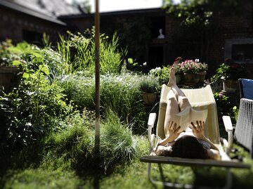 Eine Frau liegt auf einem Liegestuhl im Garten und liest ein Buch | © GettyImages/suteishi