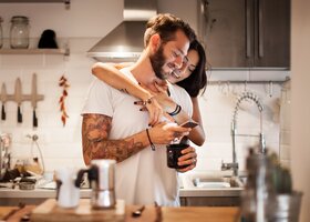 Frau umarmt ihren Partner liebevoll in der Küche, während er lächelnd auf sein Handy guckt. 