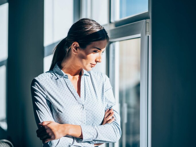 Frau steht am sonnigen Bürofenster | © Getty Images/VioletaStoimenova