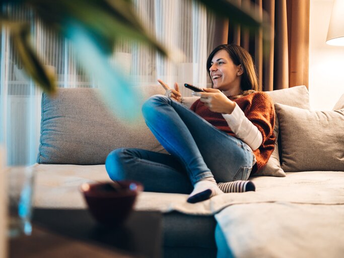 Frau guckt gut gelaunt Fernsehen auf der Couch | © Getty Images/	urbazon