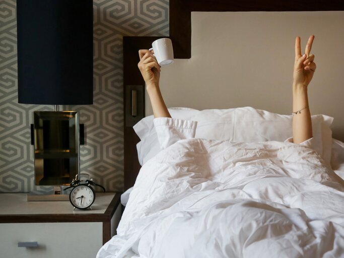 Frau liegt unter dem Bett und hält eine Tasse Kaffee in der Hand | © Getty Images/	triocean