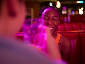 Frau sitzt bei einem Date in der Bar und lacht herzhaft, im Vordergrund verschwommen ihr Date mit einem Glas in der Hand.  | © Getty Images/Klaus Vedfelt