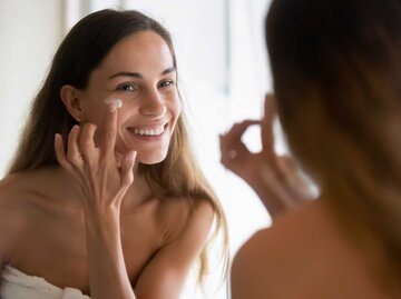 Frau trägt Gesichtscreme auf und lächelt im Spiegel | © Adobe Stock/fizkes
