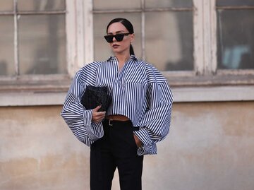 Stilbewusste Frau mit Sonnenbrille trägt gestreifte Bluse mit Puffärmeln und hält schwarze Tasche vor verwittertem Fenster. | © Getty Images/Jeremy Moeller 