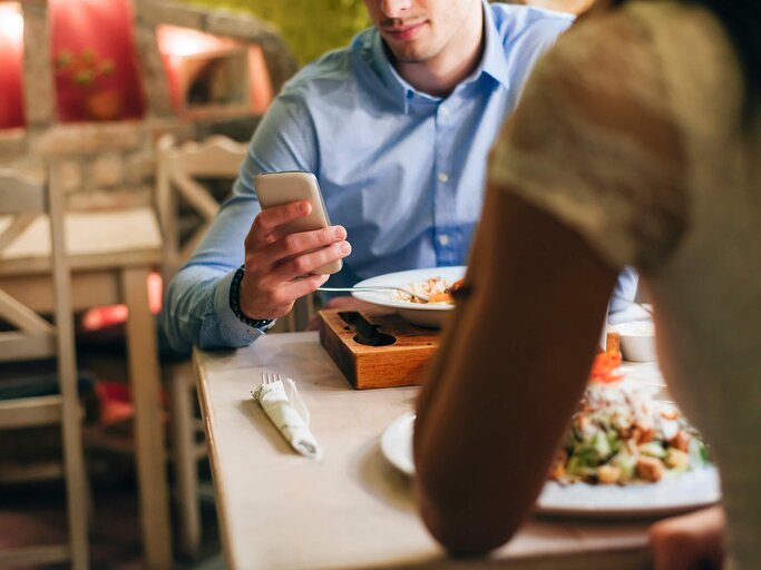 Close-up von einer Hand, die beim Essen zu zweit ein Handy hält | © gettyimages.de | Westend61
