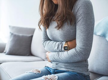 Junge Frau mit grauem Langarmshirt sitzt auf Sofa und hält sich ihren schmerzenden Bauch | © iStock | dragana991