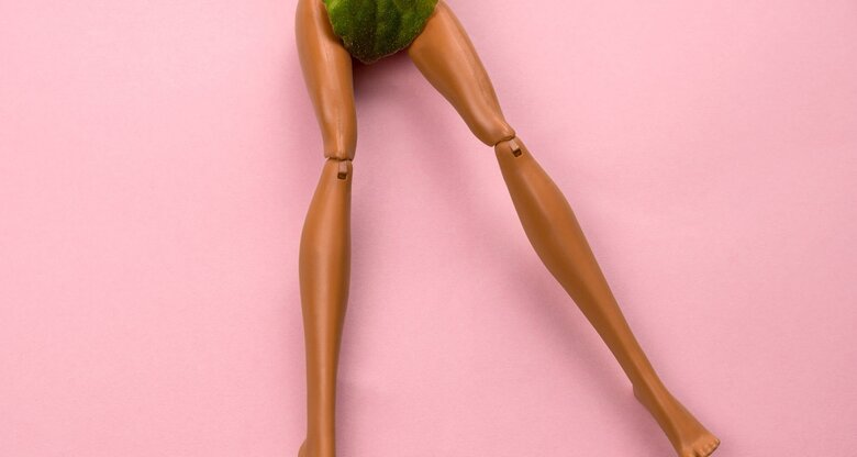 Beine einer Puppe auf Rosa Hintergrund, ein grünes Blatt bedeckt den Schambereich. | © iStock.com / dvulikaia