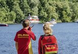Zwei Mitglieder der DLRG stehen mit Mikrofon am Seerand. | © DLRG
