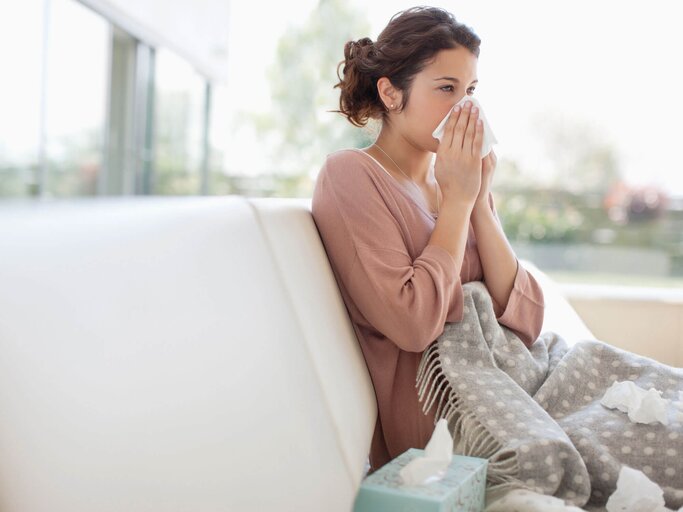 Frau sitzt mit einer Decke auf der Couch und putzt sich wegen Schnupfen die Nase. | © gettyimages.de / Paul Bradbury