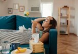 Frau liegt mit einer Erkältung auf der Couch, fühlt mit der Hand die Stirn und hält ein Fieberthermometer.  | © gettyimages.de / PixelsEffect